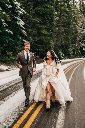 Laughing bride in white gown and UGG boots runs with groom along snowy tree-lined road.