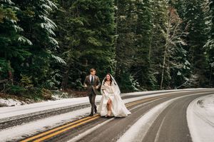Bride and groom walking on snowy mountain road surrounded by evergreen forest.