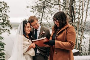 Bride, groom, and officiant reviewing wedding ceremony book outdoors in snowy forest.