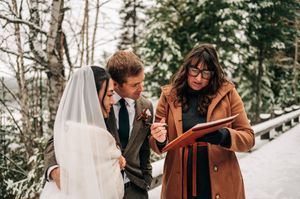 Officiant in brown coat reviews documents with bride and groom during intimate snowy winter ceremony.