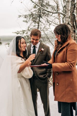 Bride reads vows while groom and officiant smile during intimate winter wedding ceremony outdoors.