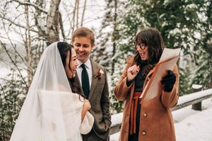 Bride and groom laugh with officiant holding folder during winter wedding ceremony in snowy forest.