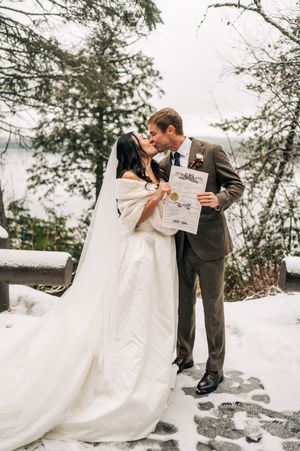 Newlyweds kiss while holding marriage certificate surrounded by snow-covered trees after ceremony.