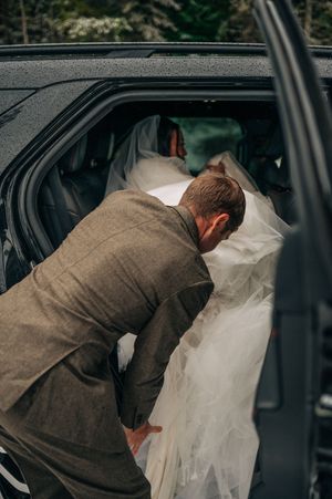 Groom in gray suit helps arrange bride's white dress and veil while getting into a dark SUV.