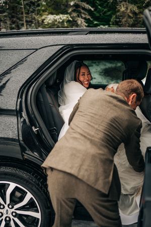 Smiling bride in white dress and veil sits in black SUV as groom leans in on rainy wedding day.