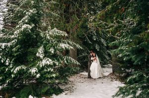 Bride and groom embrace among snow-covered evergreen trees in a serene winter forest setting.