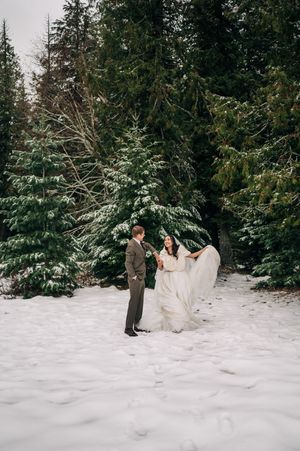 Bride twirls her flowing white gown in a snowy clearing surrounded by snow-covered evergreen trees.