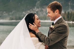 Bride and groom laugh joyfully together by a misty mountain lake on their wedding day.