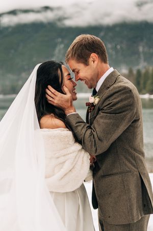 Bride and groom share a romantic moment by a snowy mountain lake, touching foreheads and smiling.