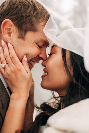 A bride and groom share a romantic moment under a wedding veil, smiling and touching foreheads, with an engagement ring visible.