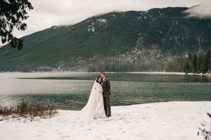 Bride and groom embrace on snowy lakeshore with misty mountain and turquoise water backdrop.
