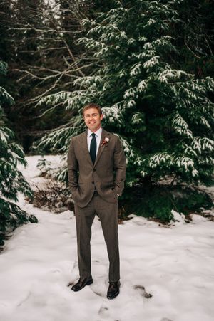 Dapper groom in brown suit with boutonniere stands confidently before snow-covered evergreen trees.