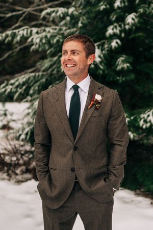 Joyful groom in brown tweed suit with boutonniere smiles outdoors in snowy winter setting.