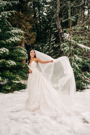 Bride in strapless white gown tosses her veil in a snowy evergreen forest on her wedding day.