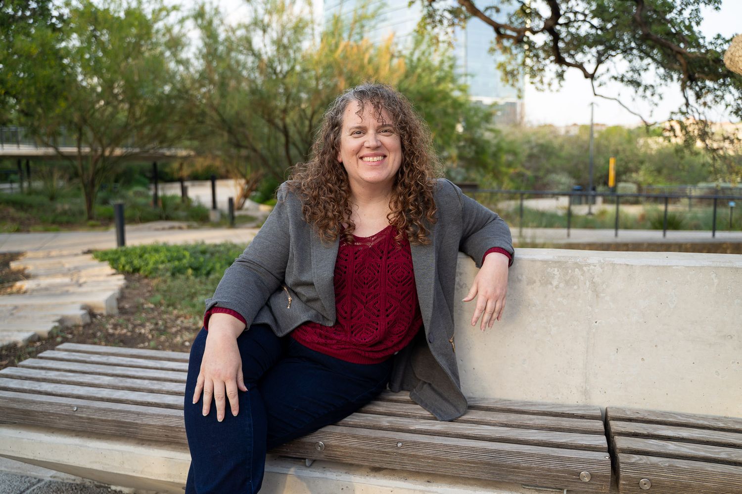 Woman with curly hair smiling on a park bench, wearing a gray blazer and burgundy top outdoors.