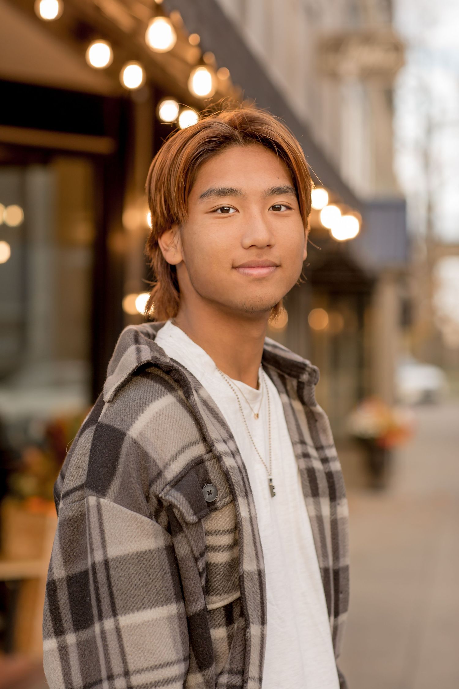 Young Asian man with highlighted hair wearing plaid shirt standing on urban city street.