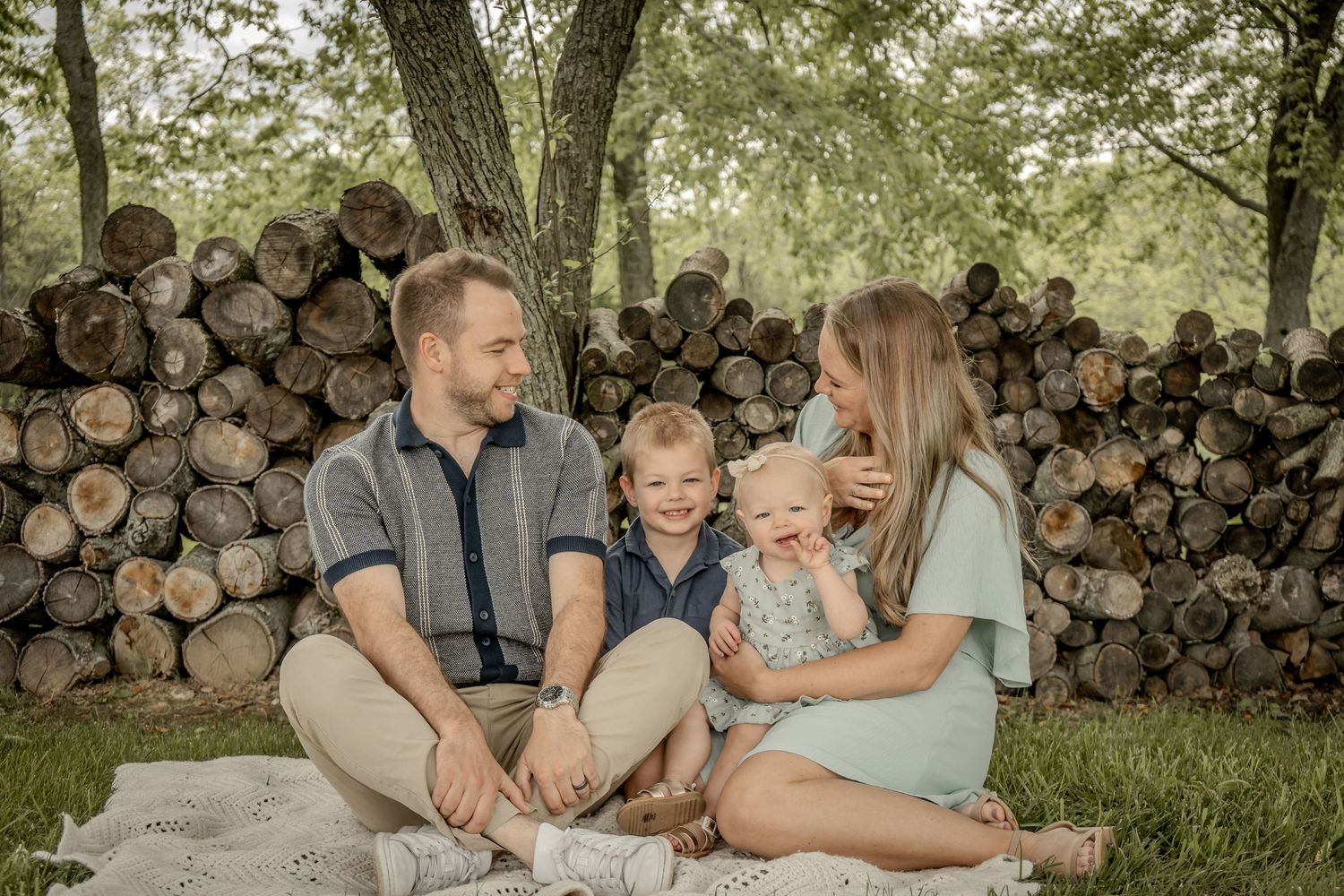 Happy young family of four sitting on blanket outdoors near stacked logs in park.
