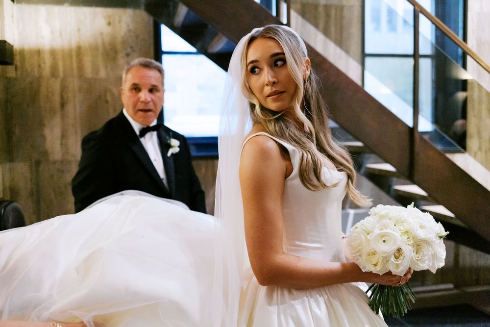 Bride in white gown holding white floral bouquet with father of the bride behind her near staircase.