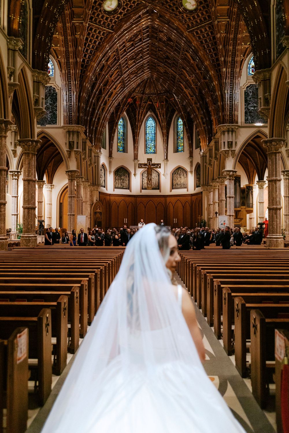 Bride in white gown and veil walks down the aisle of the grand Holy Name Cathedral during Catholic wedding ceremony.