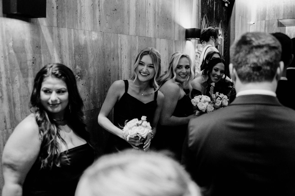 Bridesmaids in black dresses smiling candidly aand holding bouquets in cathedral vestibule.