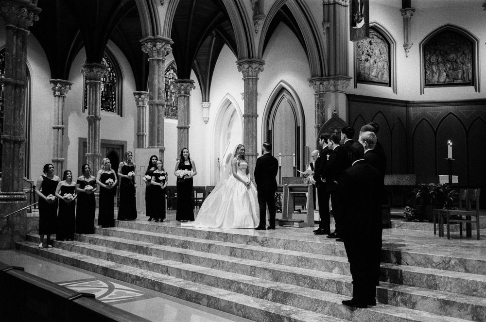 Black and white photo of a bride and groom at the altar of a grand church with bridesmaids standing on steps.