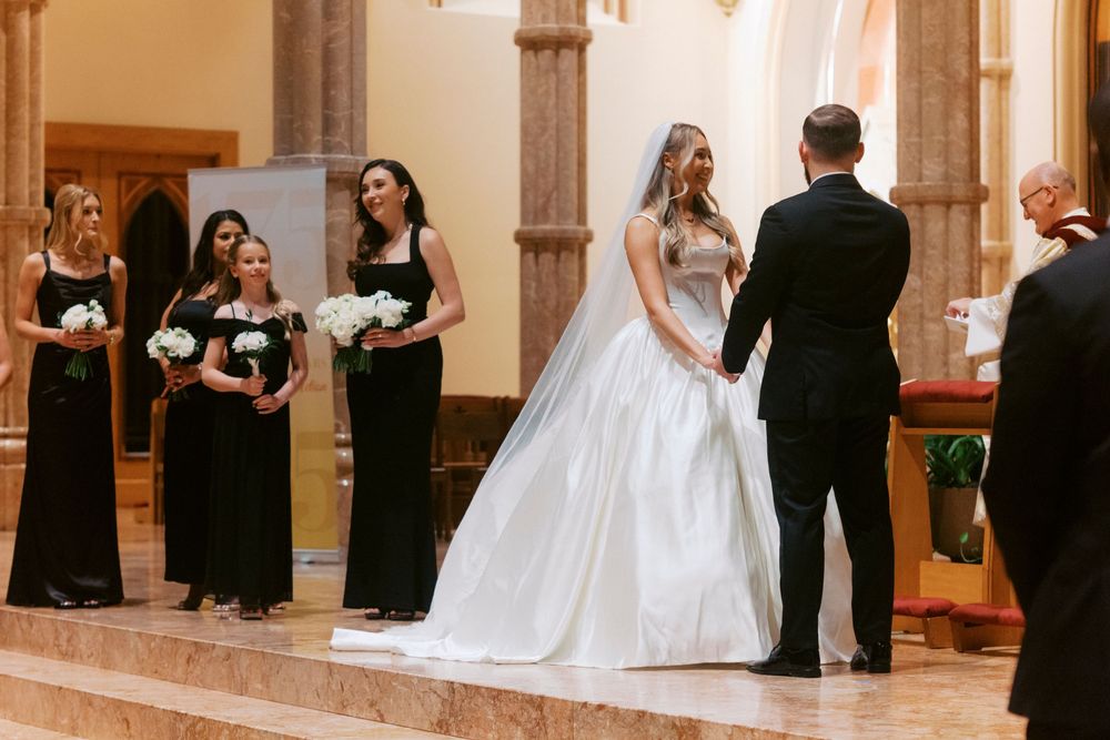 Bride in white ball gown stands at church altar with bridesmaids in black dresses holding white bouquets during wedding ceremony.