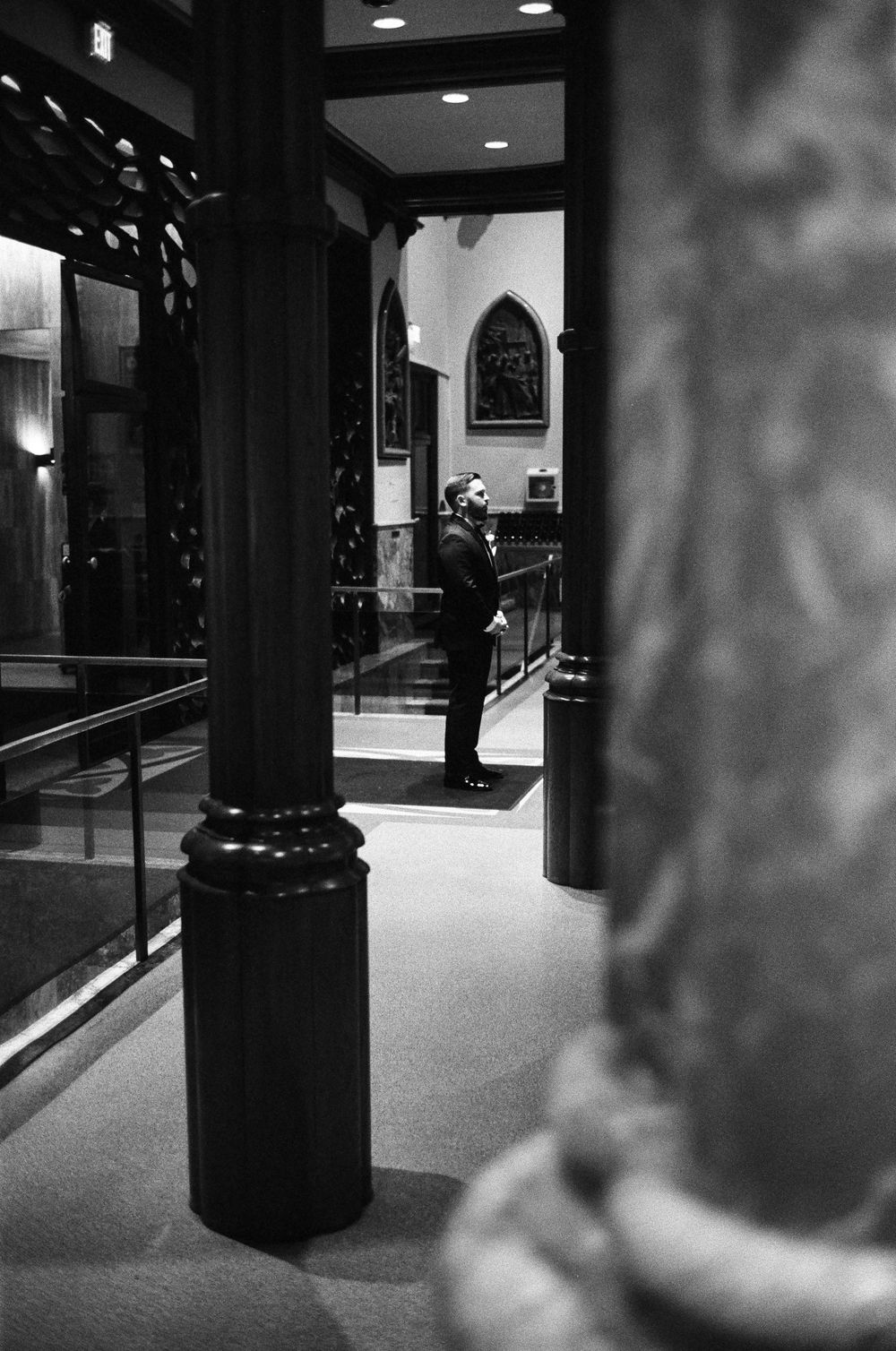 Groom stands alone in church corridor before wedding ceremony in dramatic black and white photo.