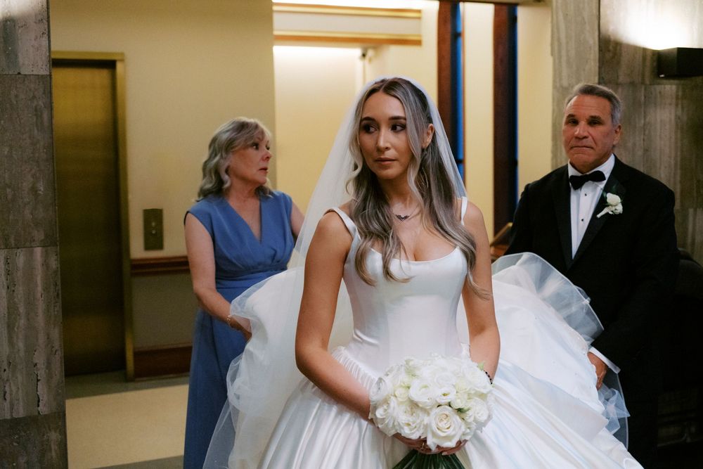 A bride in a white ballgown holds a white bouquet while her parents stand behind her before the Catholic ceremony.