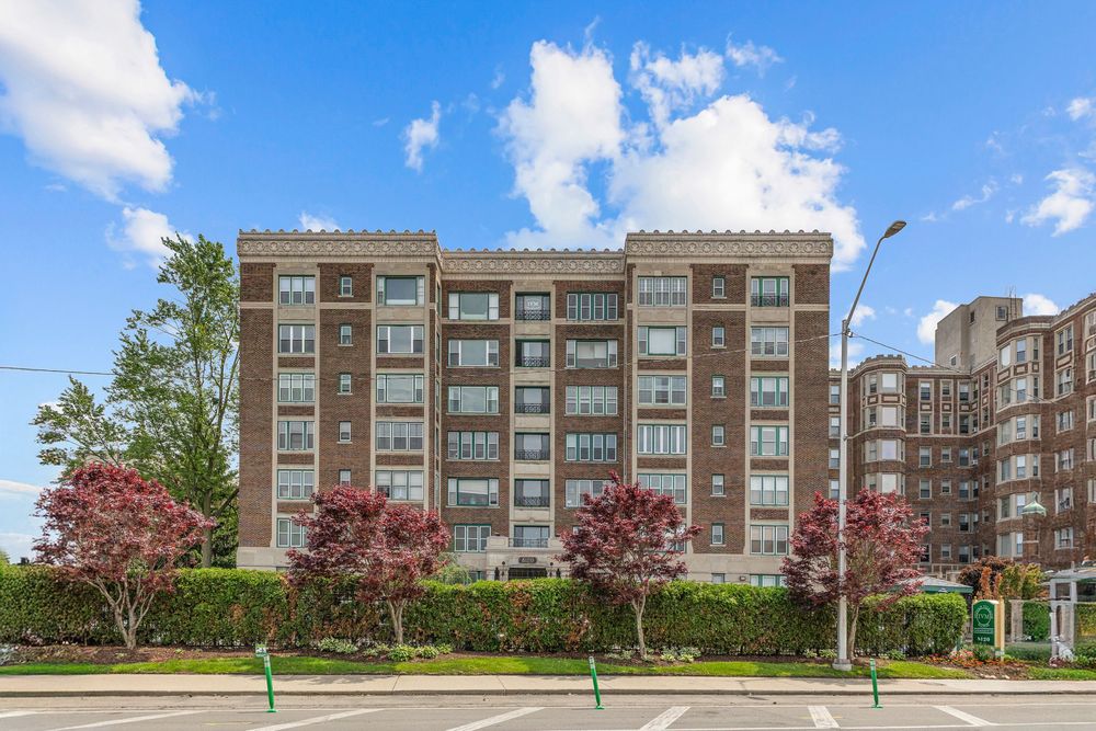 Brick mid-rise apartment building with balconies, red maple trees, and manicured hedges under a bright blue sky. Detroit Michigan