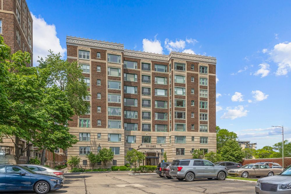 Brick mid-rise condo building with balconies, surrounded by mature trees and a parking lot under a bright blue sky. Detroit Michigan