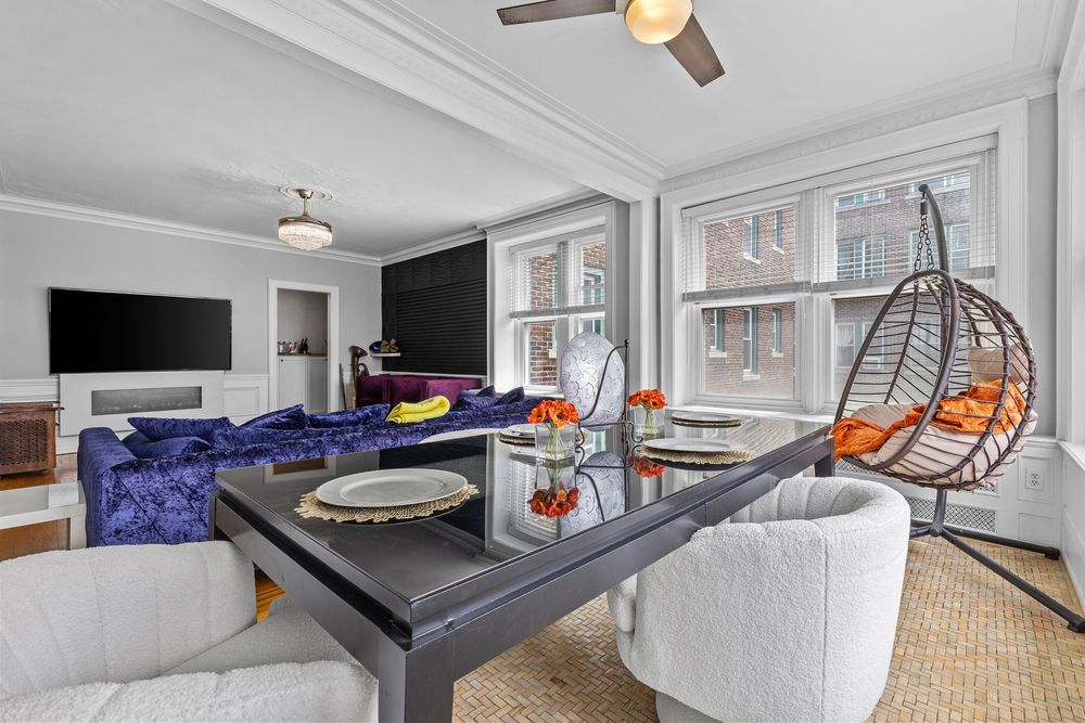 Bright dining room with glass table, hanging egg chair, crown molding, ceiling fan, and blue velvet sofa in background. Detroit Michigan