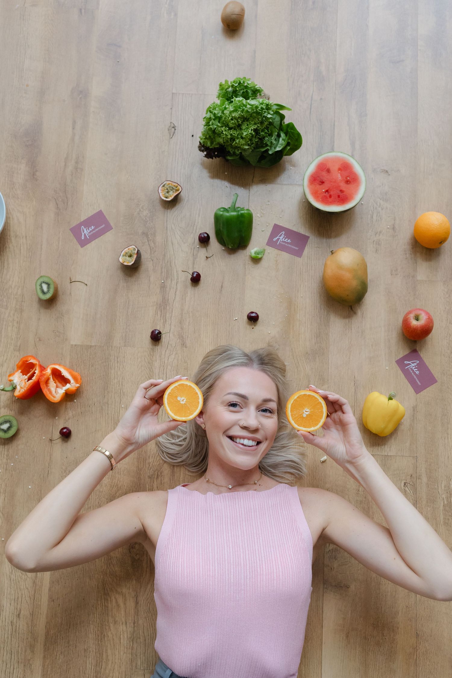 Smiling blonde woman lying on wooden floor holding orange halves surrounded by fresh fruits and vegetables.