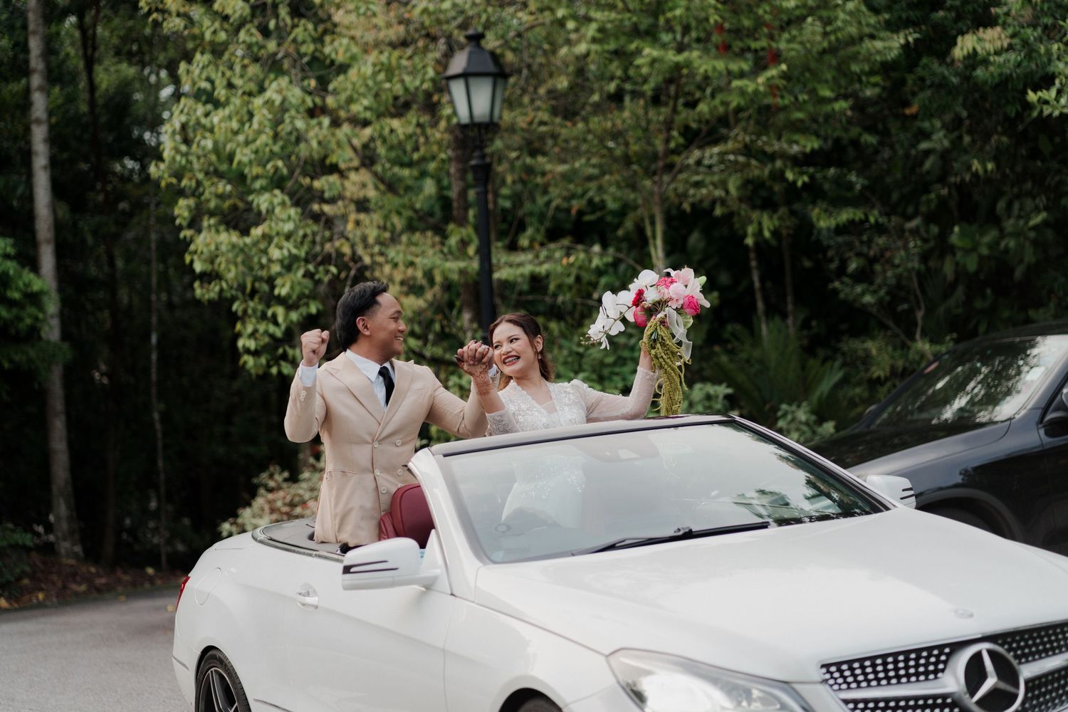 Newlywed couple celebrating in a white Mercedes convertible, bride holding pink bouquet, groom in beige suit, lush green trees.