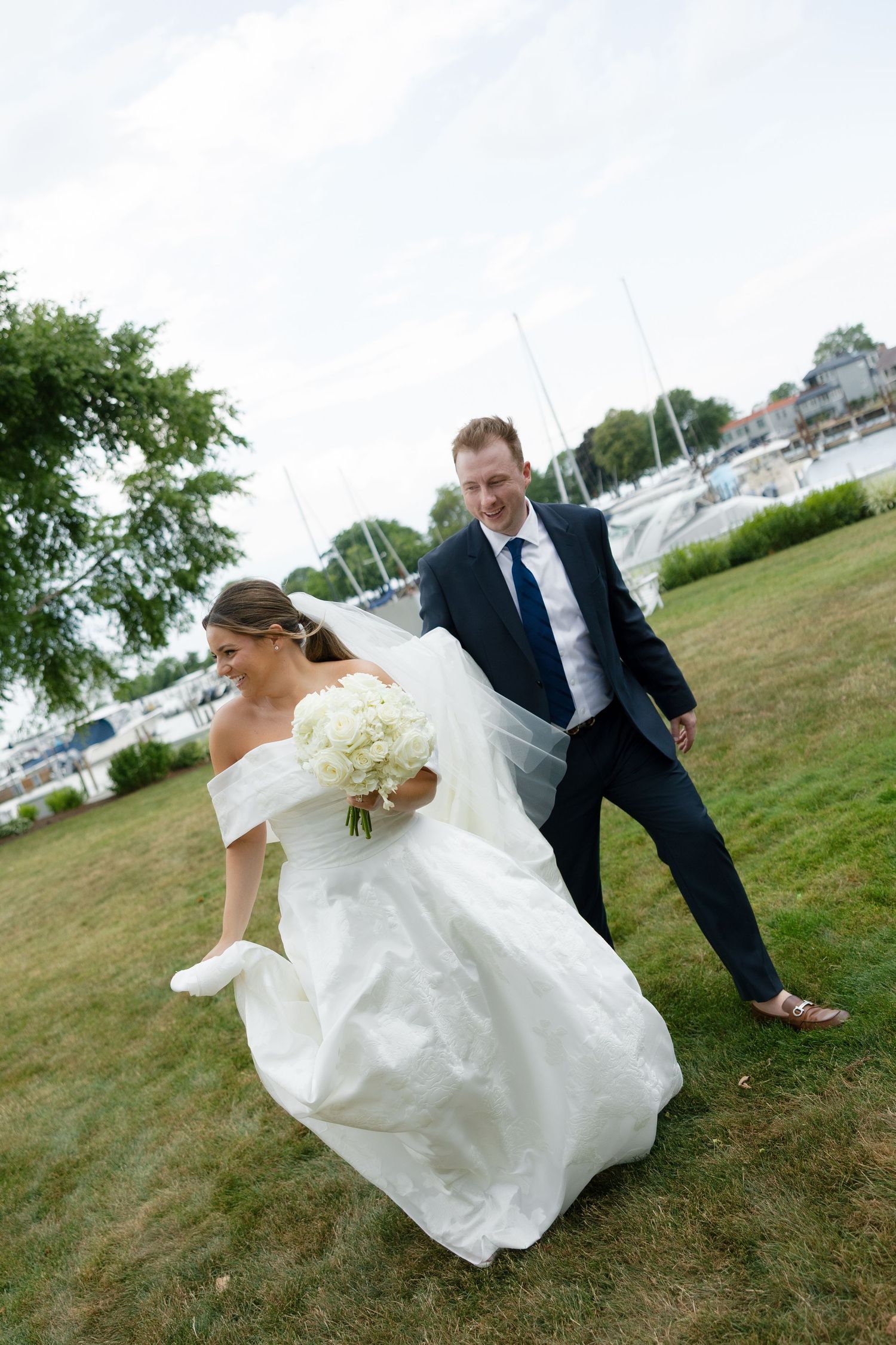 Bride and groom walk across green lawn near marina with sailboats in background.