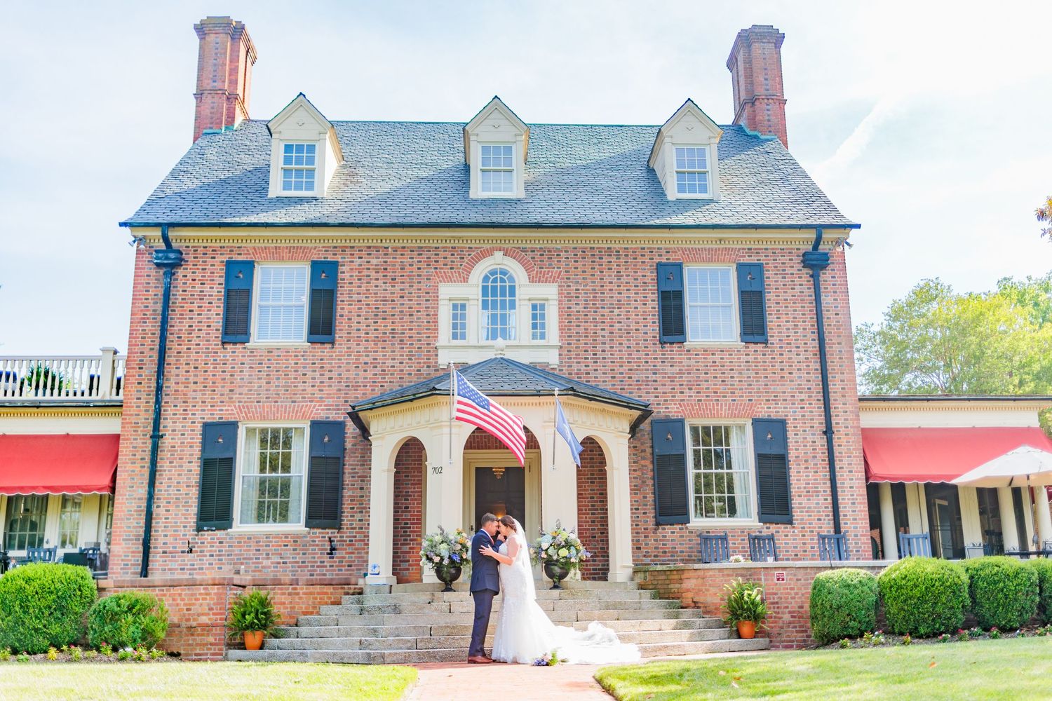 Wedding couple posing in front of elegant brick colonial mansion with white columns.