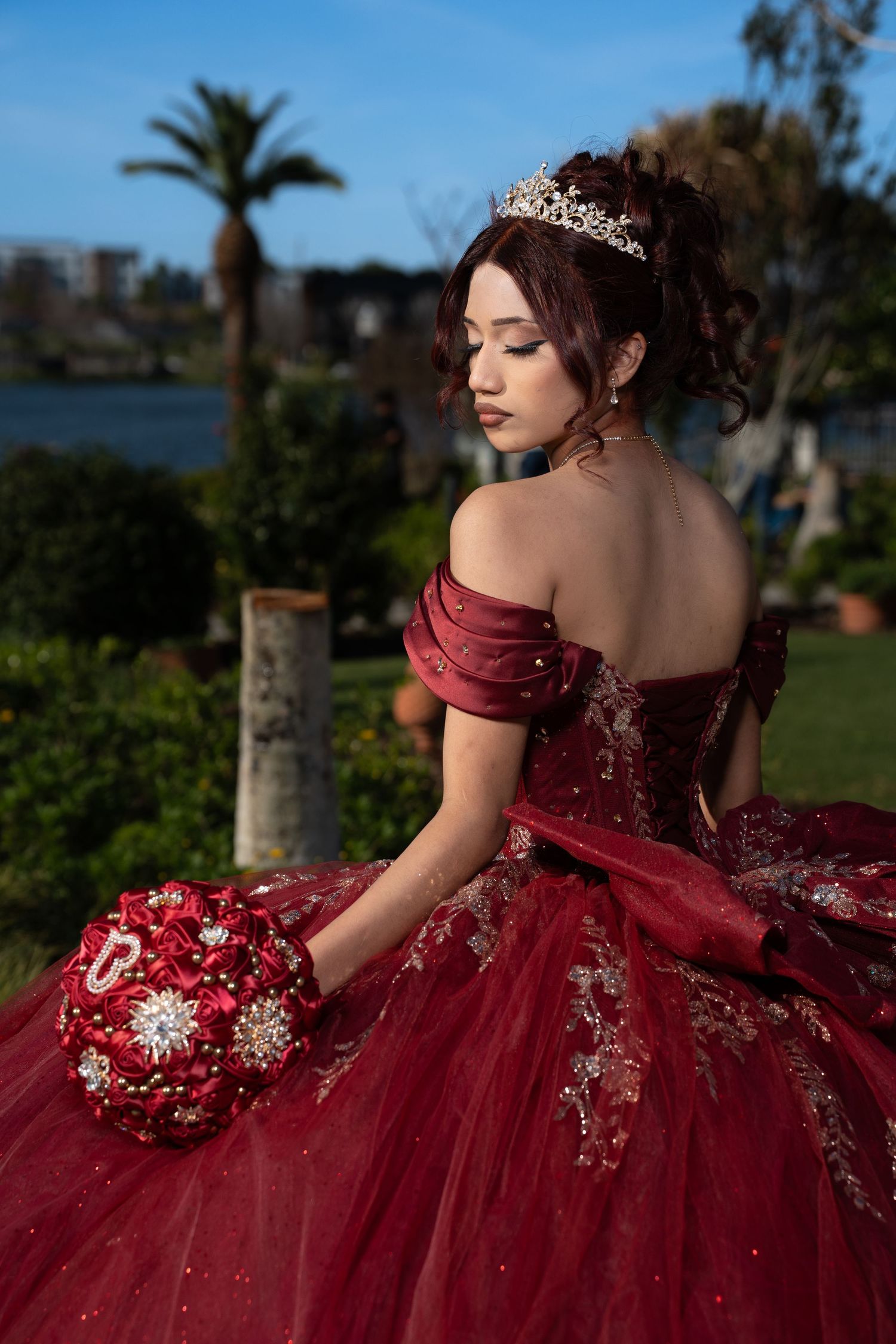 Young woman in red off-shoulder quinceañera ball gown with tiara holds floral bouquet outdoors near water.