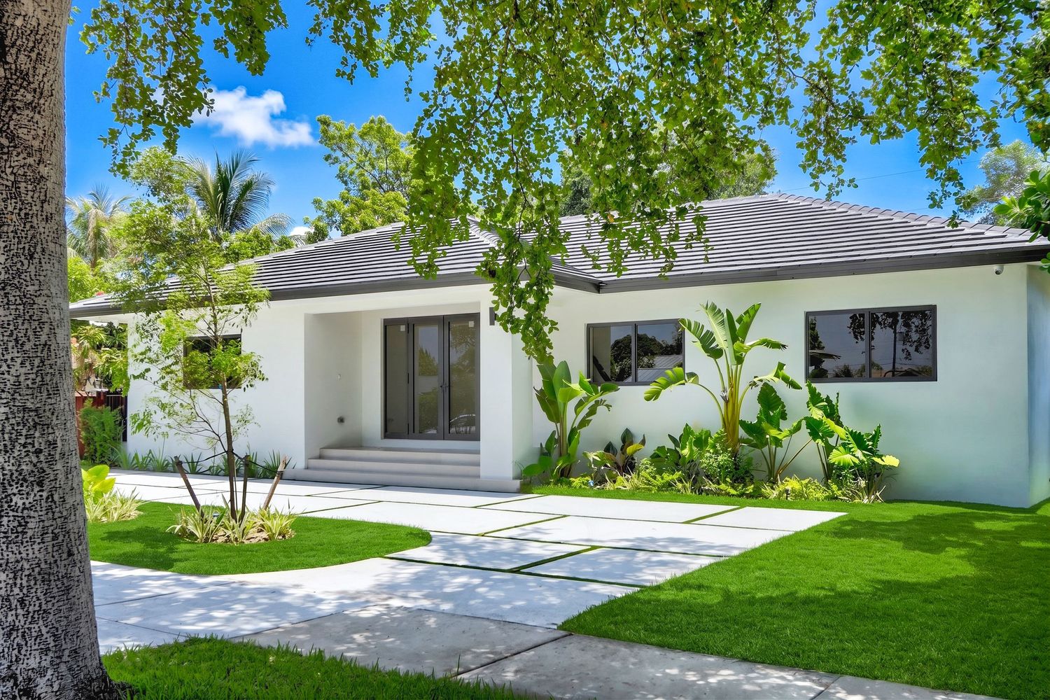 Modern white single-story home with lush green lawn, tropical landscaping, and a welcoming entrance in a sunny setting.