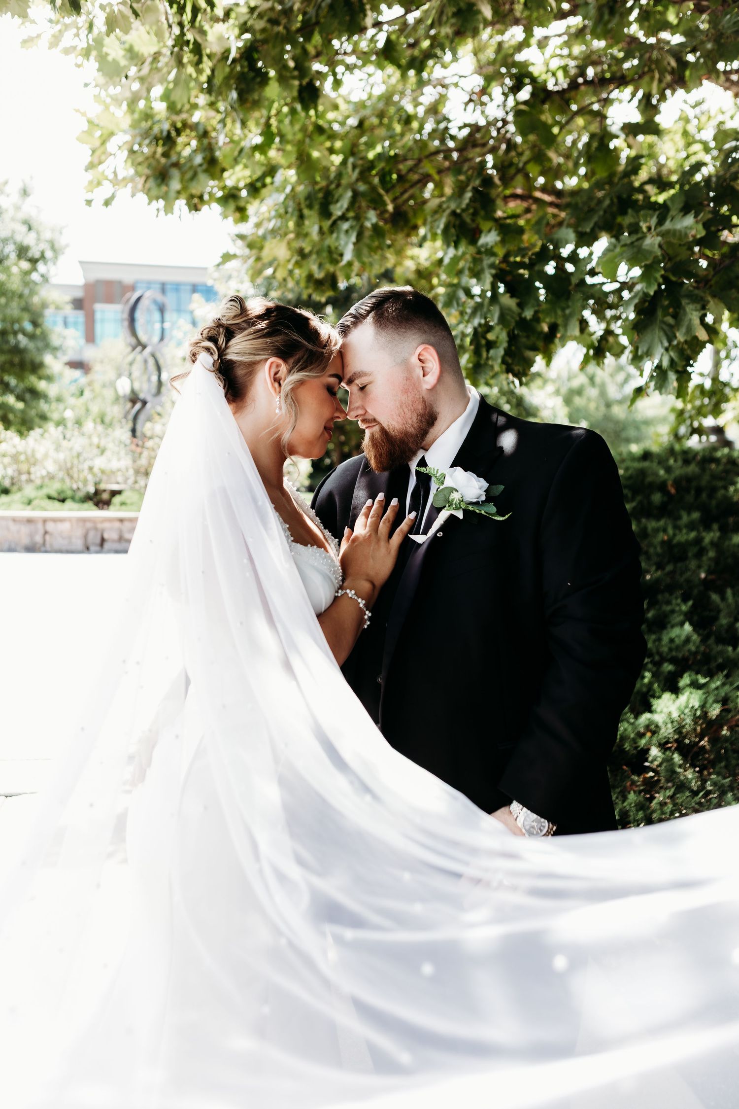 Bride and groom touching foreheads romantically under green trees, bride's veil flowing in the breeze on their wedding day.
