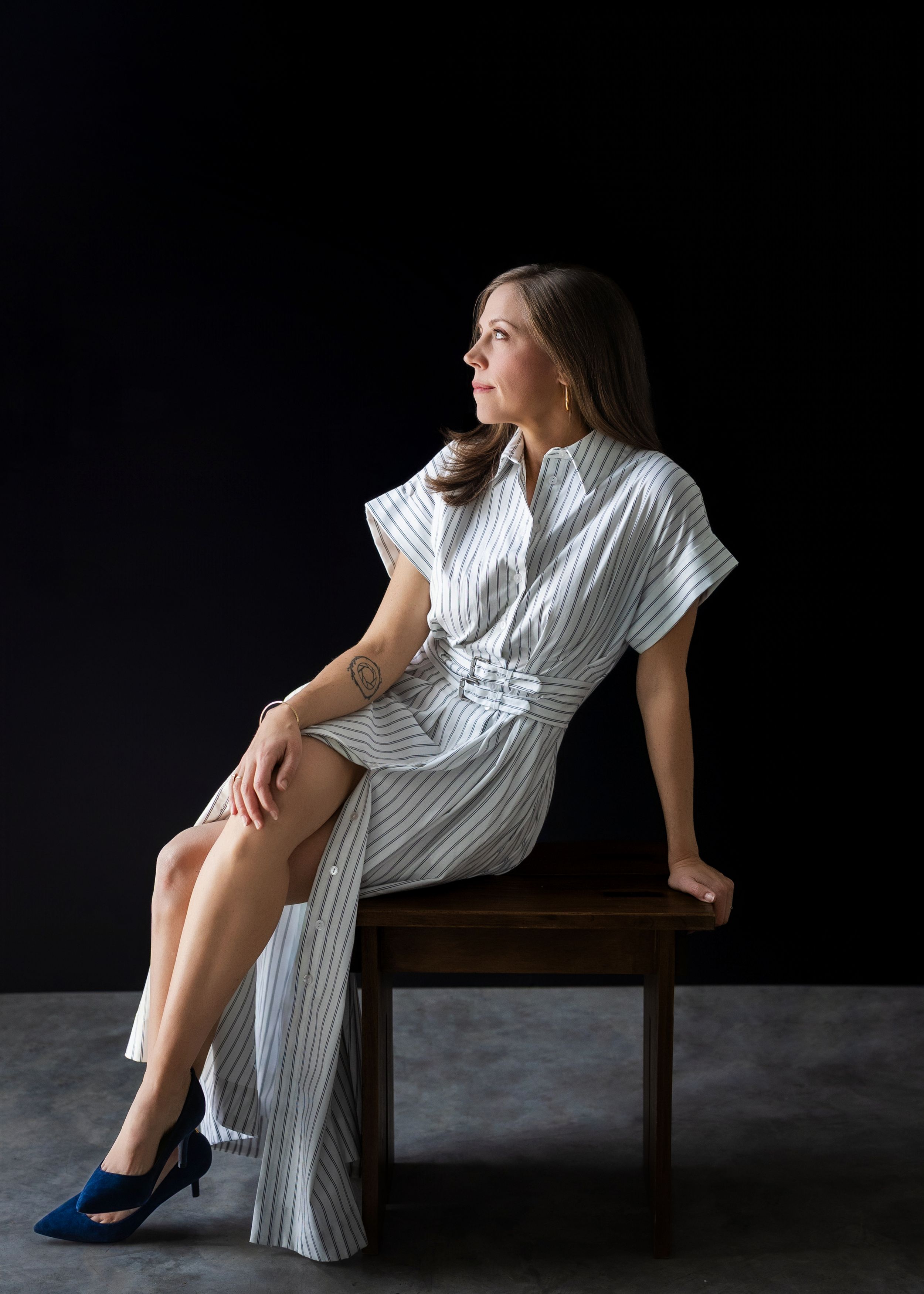 Woman in striped dress seated on wooden chair against dark background, gazing thoughtfully to the side, wearing blue heels.