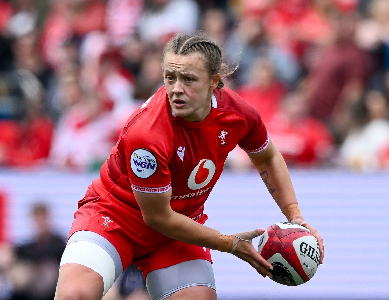 Female rugby player in red Wales jersey holding ball during a match on a sunny day.
