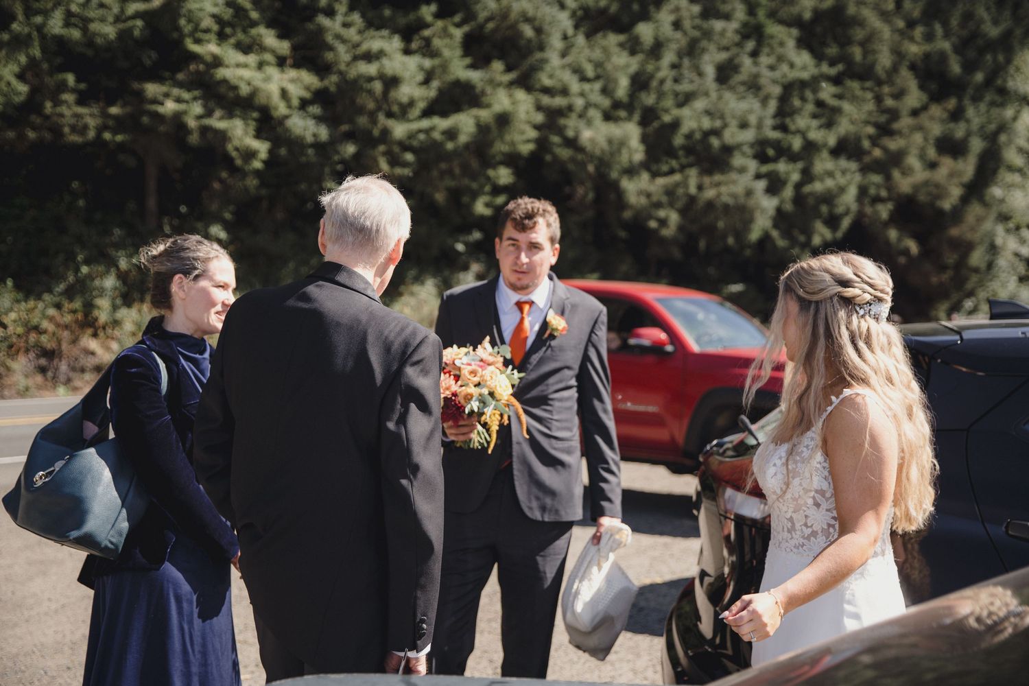 Bride and groom exchanging bouquet outdoors near trees and red car with wedding guests.
