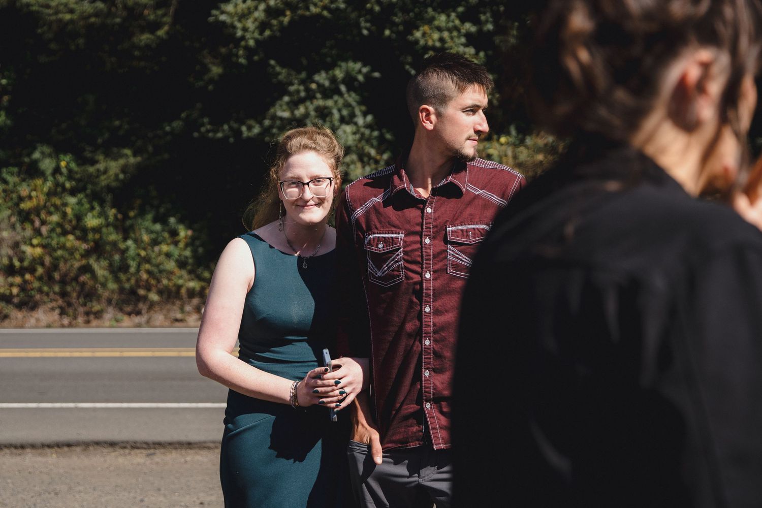 A couple in formal attire poses outdoors near a road, with a blurred figure in the foreground.