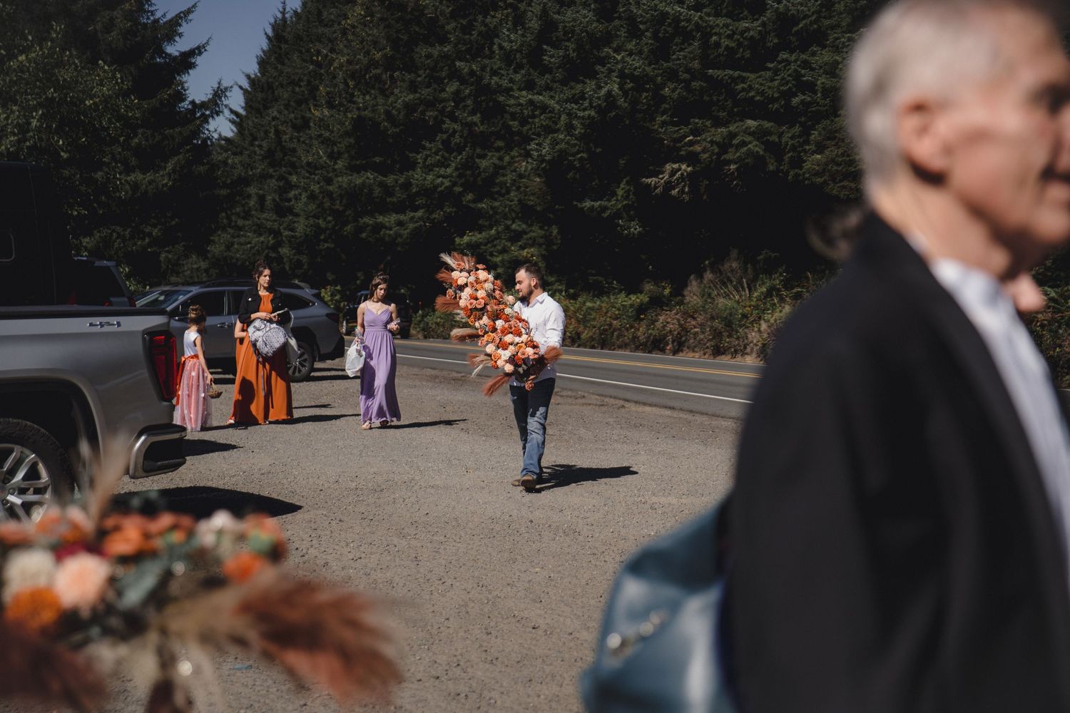 Wedding party walks across a parking lot carrying colorful floral arrangements on a sunny day near tall trees.