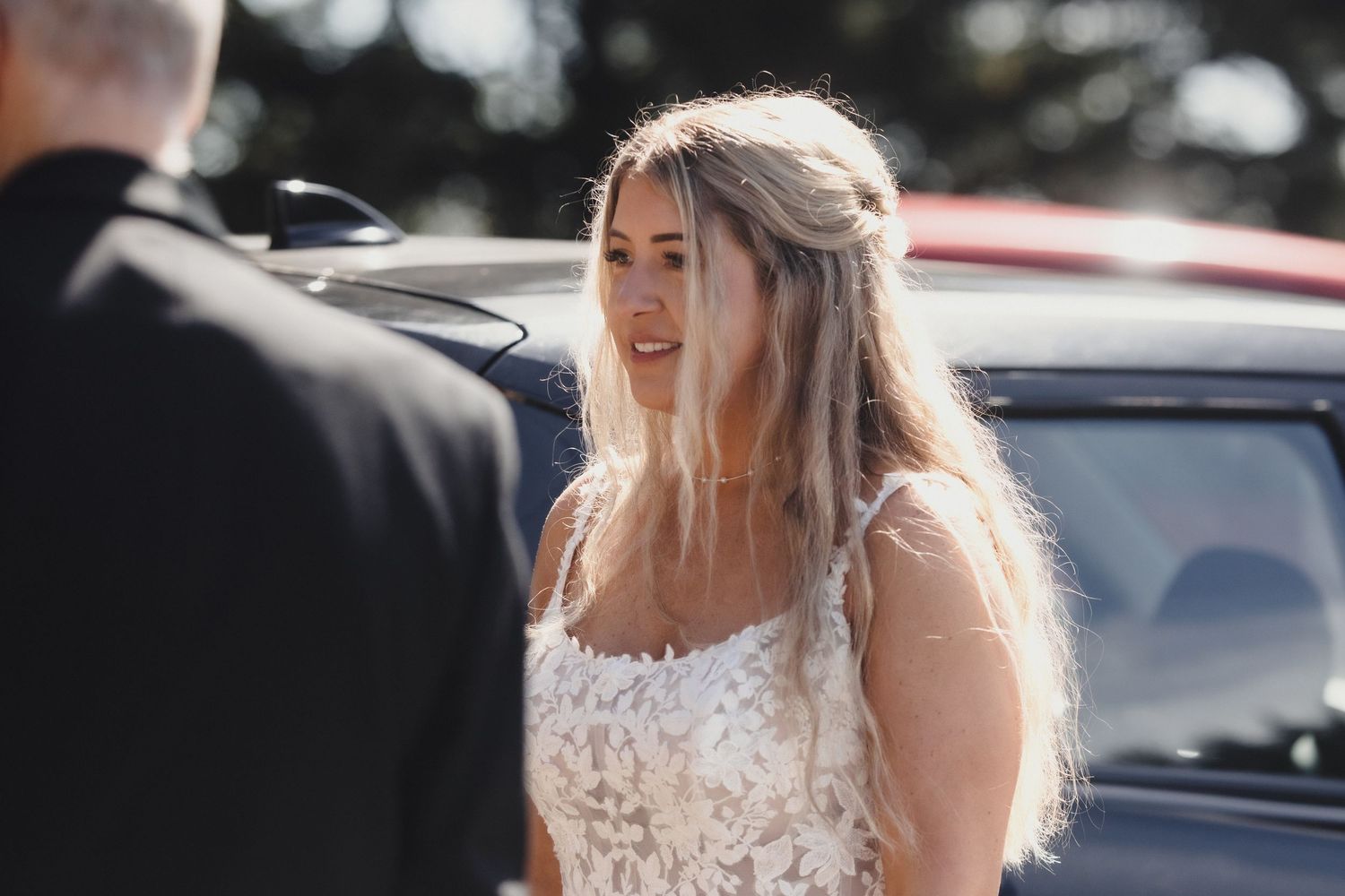 Bride in white lace dress smiling outdoors on sunny wedding day near dark car.