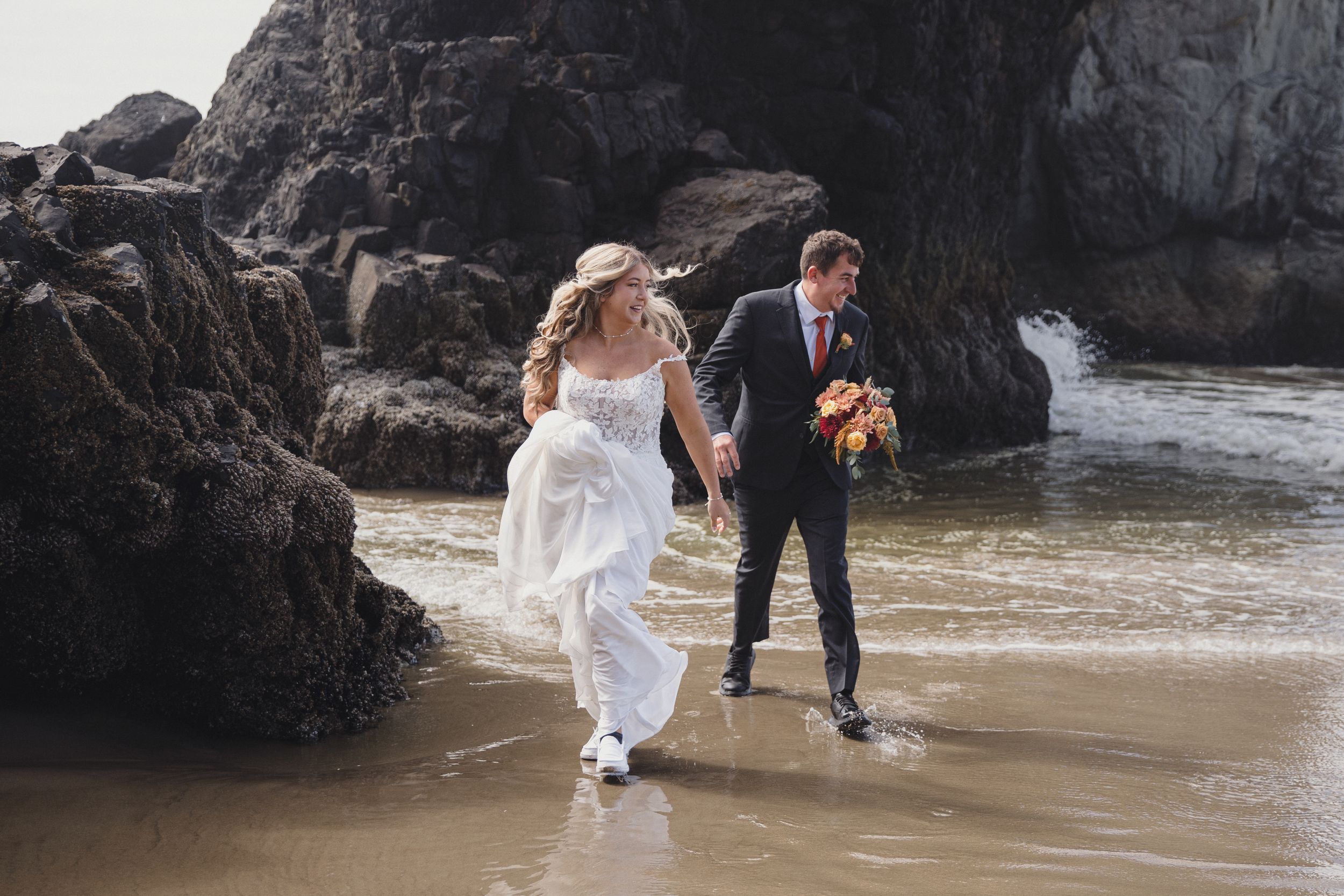 Bride and groom walking hand in hand along a rocky beach shoreline, bride holding her white dress above the water.