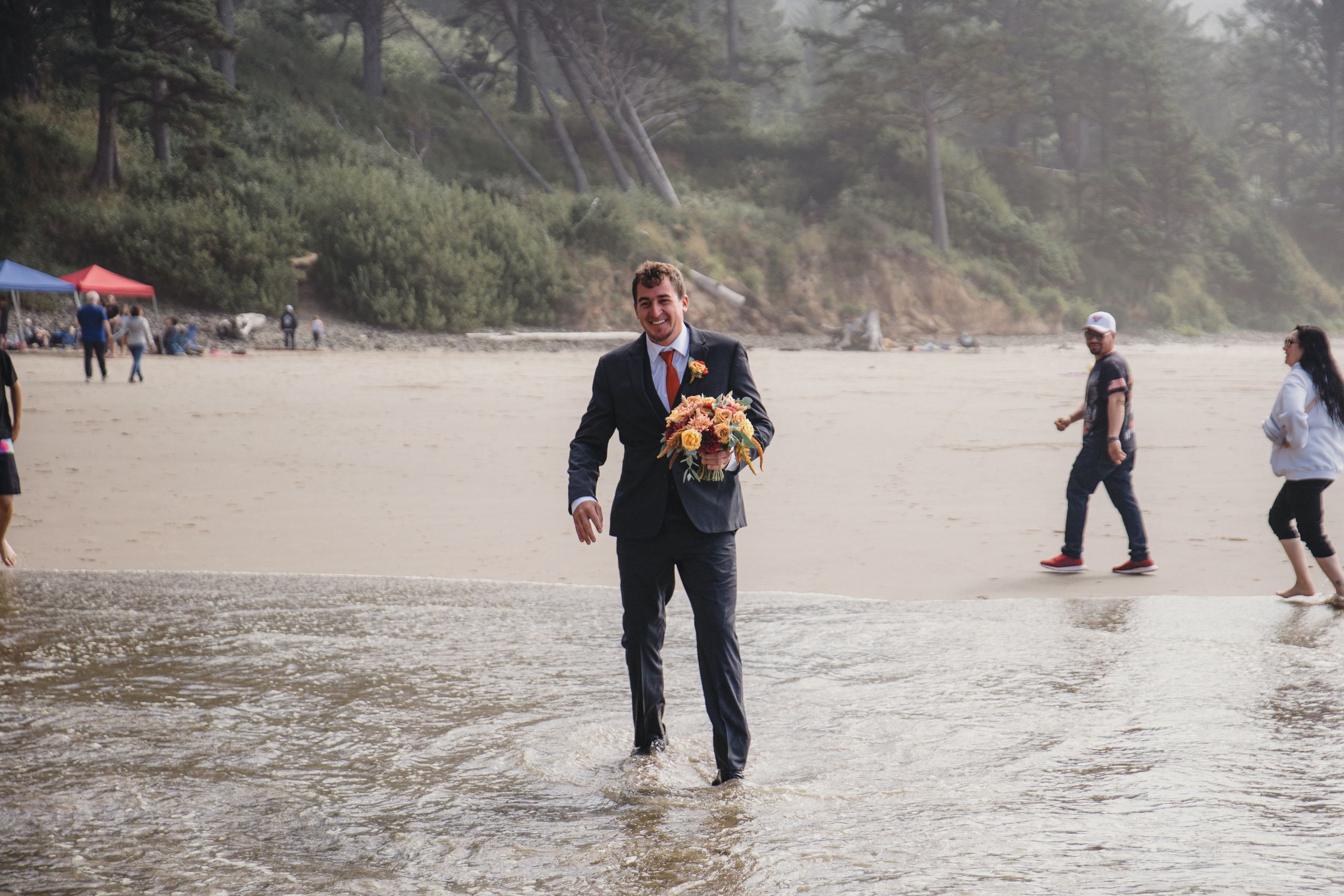 Groom in navy suit carrying bridal bouquet walking barefoot on sandy beach during outdoor wedding ceremony.