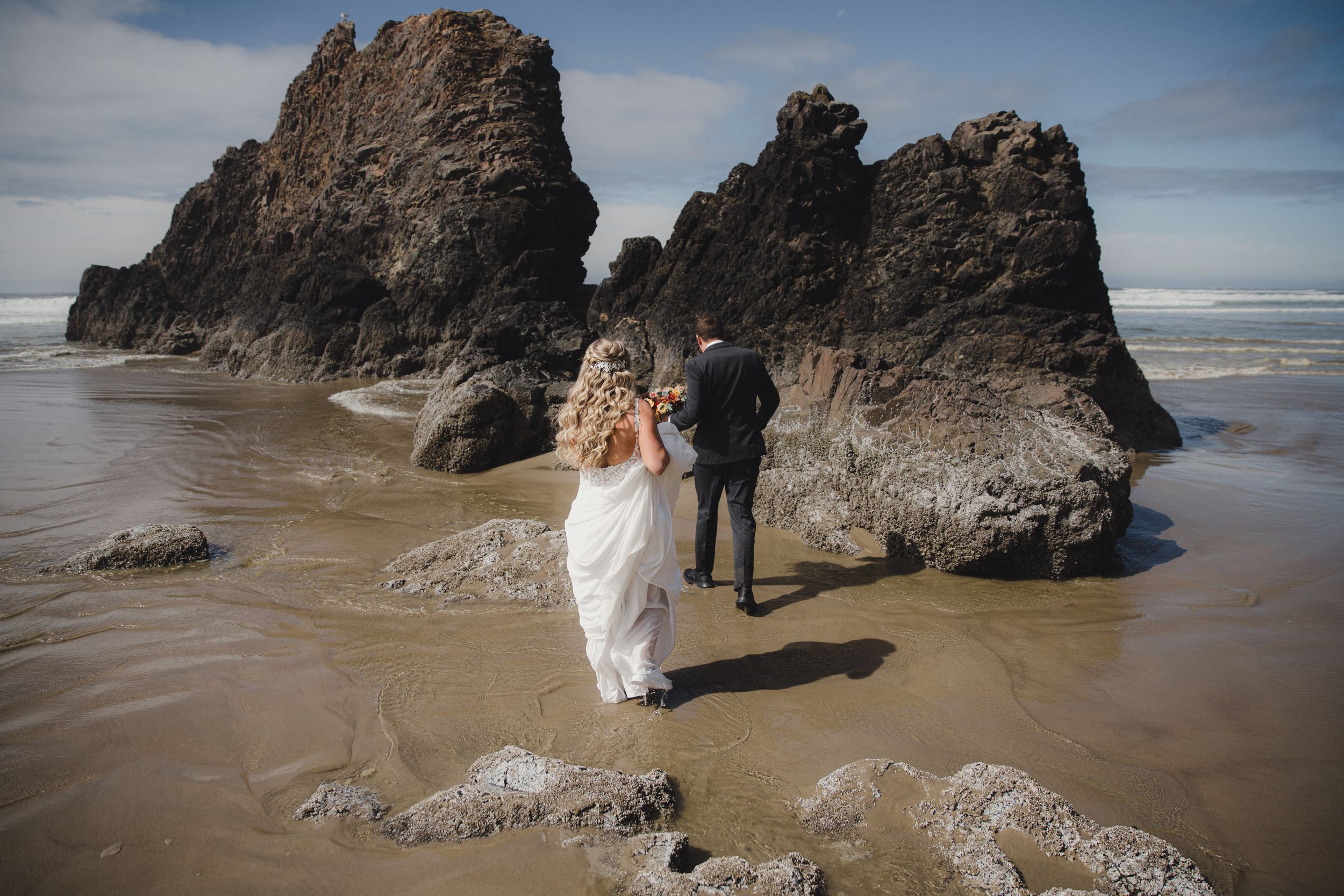 Bride and groom standing together on a sandy beach surrounded by dramatic dark rock formations by the ocean.