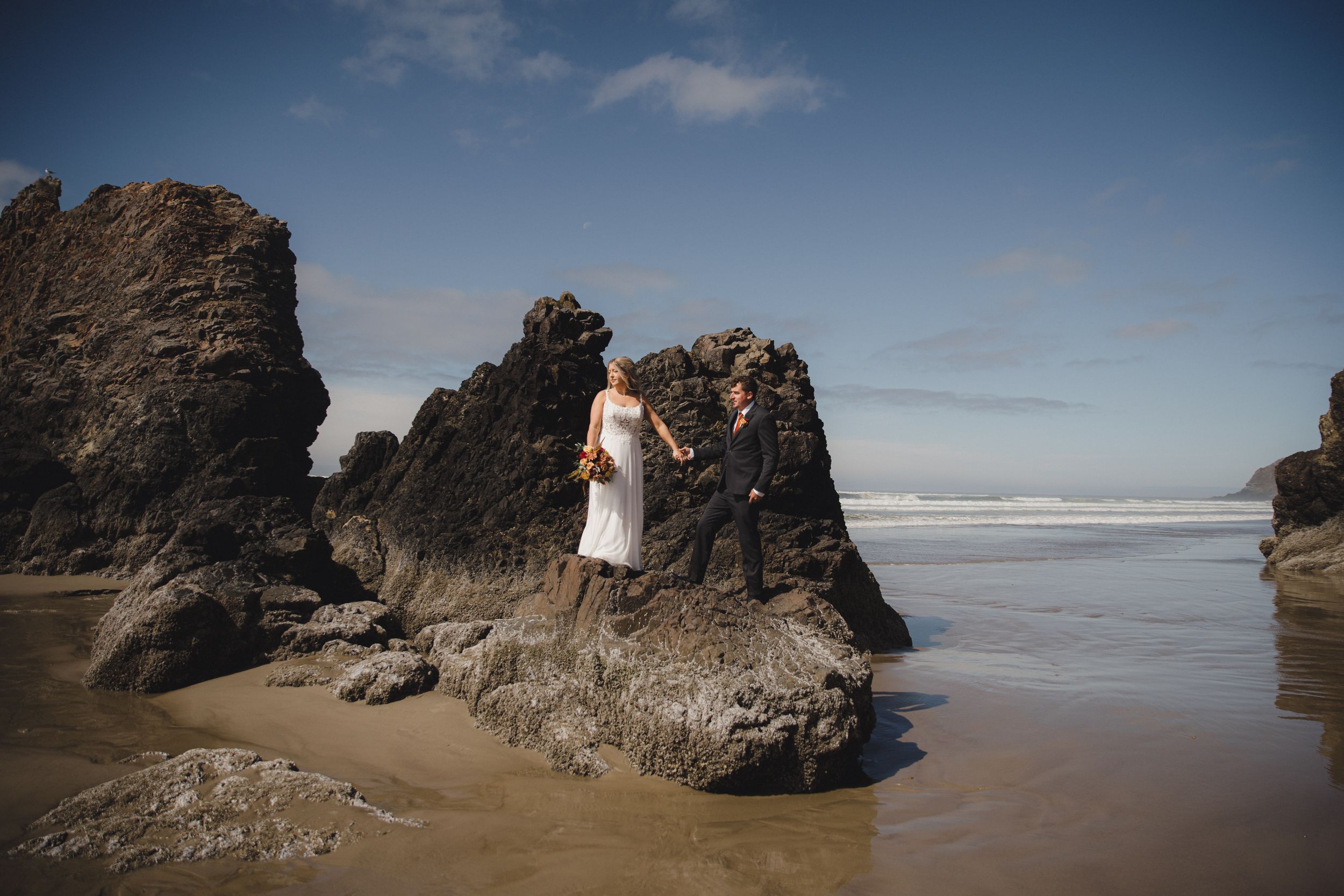 Bride and groom standing on large rocks at a scenic beach on their wedding day.
