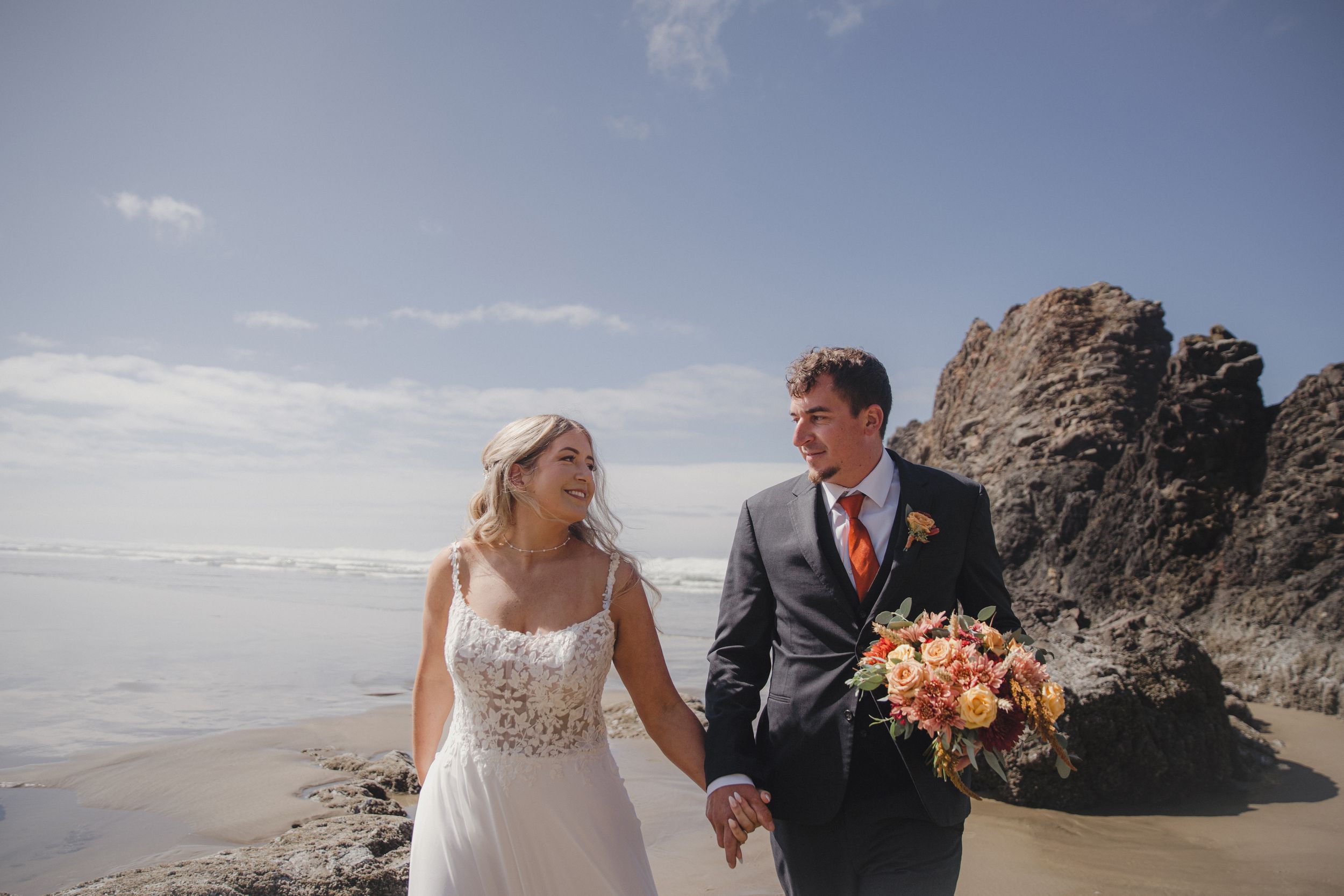 Smiling bride and groom holding hands and a bouquet on a sunny beach wedding day.