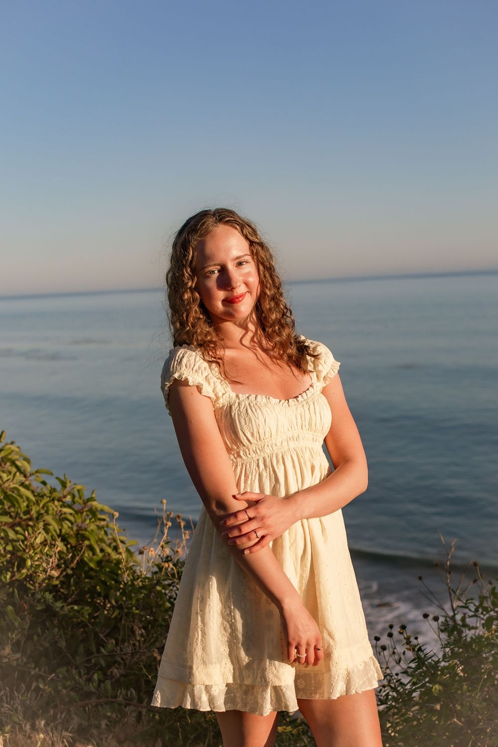Young woman in white sundress poses by ocean cliff at golden hour sunset with blue sky background.
